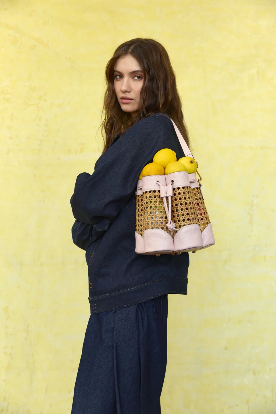 Model carrying a blush Solihiya Flower Bucket Bag over one shoulder against a yellow backdrop.