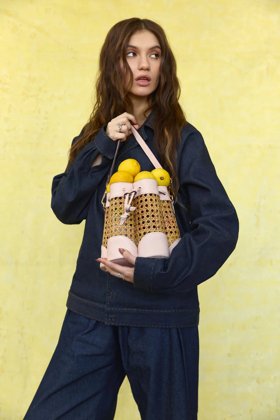 Model holding a blush Solihiya Flower Bucket Bag filled with lemons against a yellow backdrop.