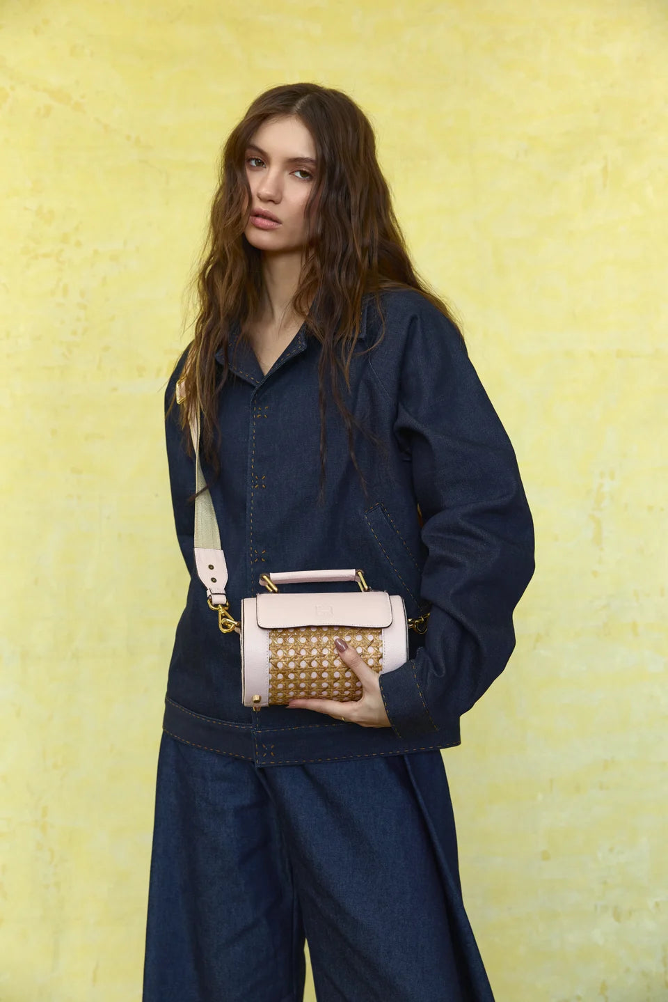 Model wearing a pale pink Solihiya Rollers Mini crossbody with a navy outfit against a yellow backdrop.